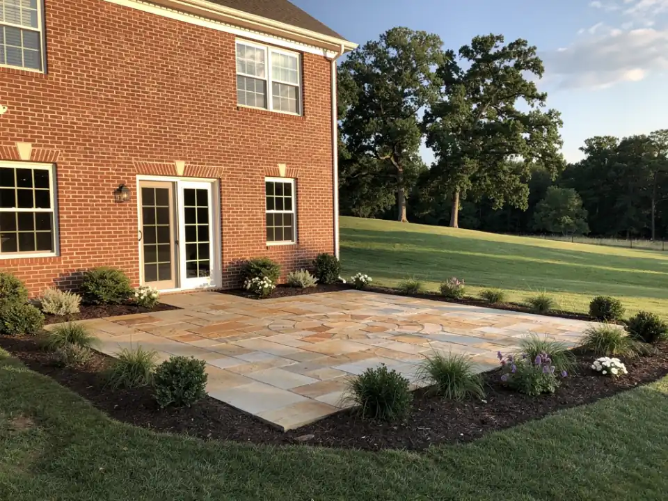 Stone paver patio and planted beds behind a brick home in late afternoon light